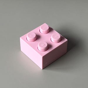 May include: A light pink building block with four raised circular studs on top. The block is square-shaped and sits on a gray surface. The block is a solid color and has a smooth texture.
