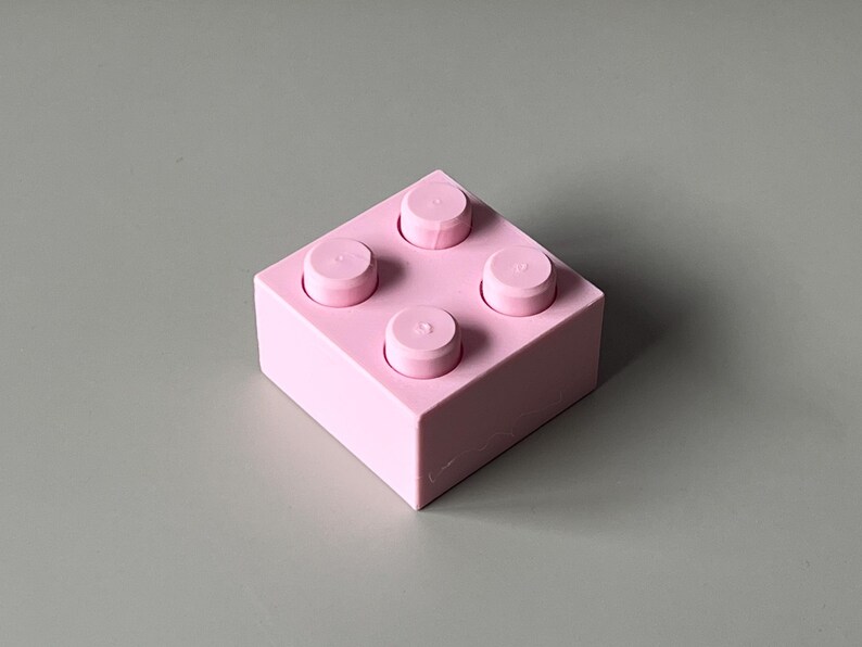 May include: A light pink building block with four raised circular studs on top. The block is square-shaped and sits on a gray surface. The block is a solid color and has a smooth texture.