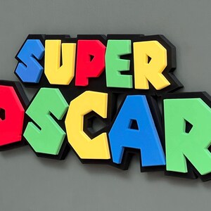 Puede incluir: Un letrero 3D colorido que dice "SUPER OSCAR". Las letras son azules, rojas, amarillas y verdes, con un contorno negro. Las letras est&aacute;n en relieve, creando un efecto 3D. El dise&ntilde;o es l&uacute;dico y recuerda a un universo de juego.