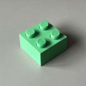 May include: A solid green building block with four raised circular studs on top. The block is square-shaped and has a smooth, matte finish. The color is a vibrant, light green.