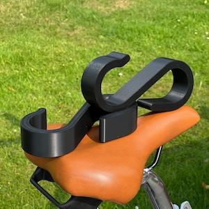 May include: A black bicycle accessory is mounted on a brown leather bicycle seat. The accessory has a curved design with two rounded ends. The seat is attached to a bicycle with a silver metal frame, set against a backdrop of green grass.