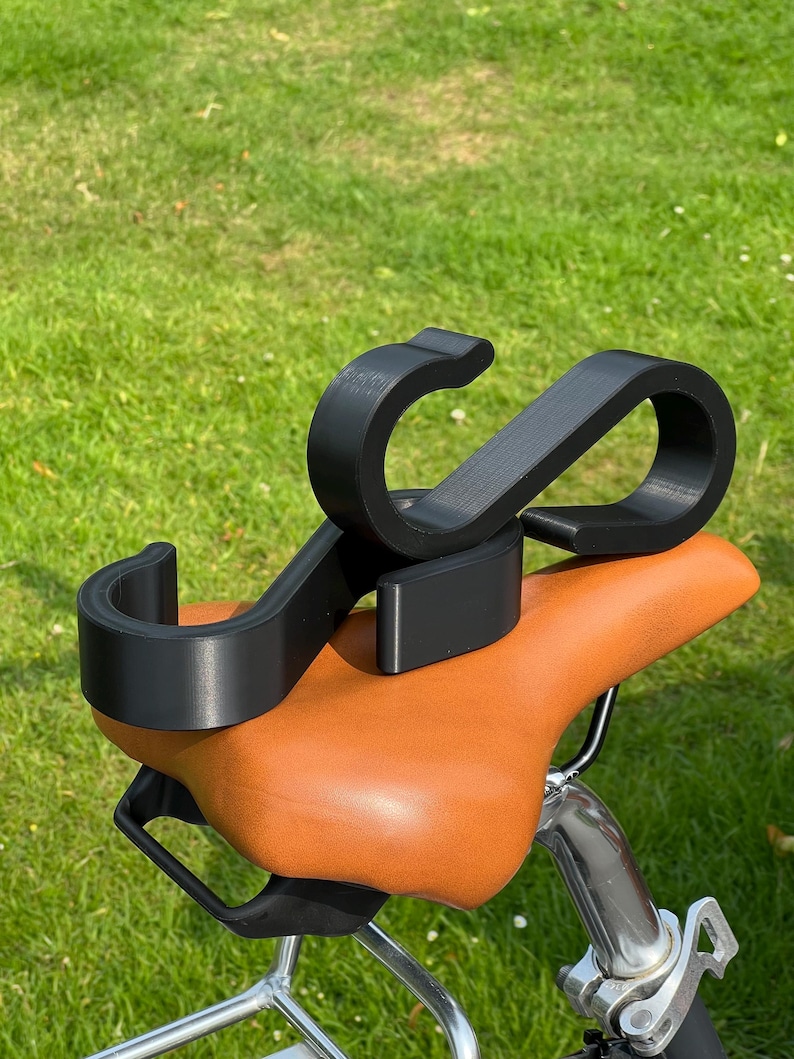 May include: A black bicycle accessory is mounted on a brown leather bicycle seat. The accessory has a curved design with two rounded ends. The seat is attached to a bicycle with a silver metal frame, set against a backdrop of green grass.
