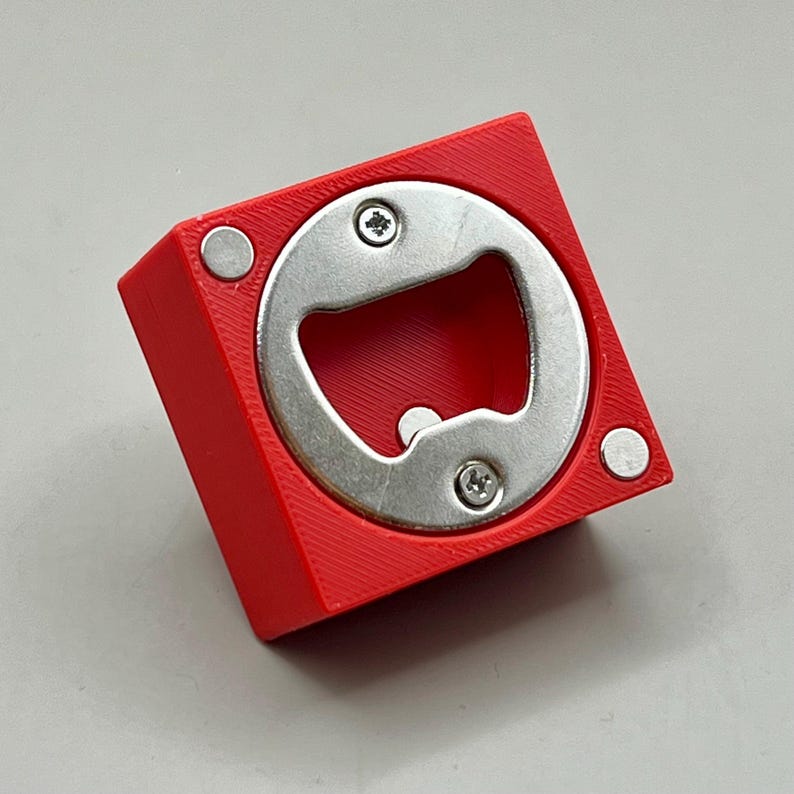 Op de afbeelding: Een rode vierkante flesopener met een zilveren metalen opener in het midden. Twee zilveren magneten zitten aan de zijkanten. De opener is vastgezet met twee schroeven.