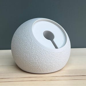 May include: A spherical white charging dock with a textured, matte exterior surface. The top features a smooth, recessed circular area with a central hole and a vertical slot extending downward, designed to hold a charging cable securely in place.