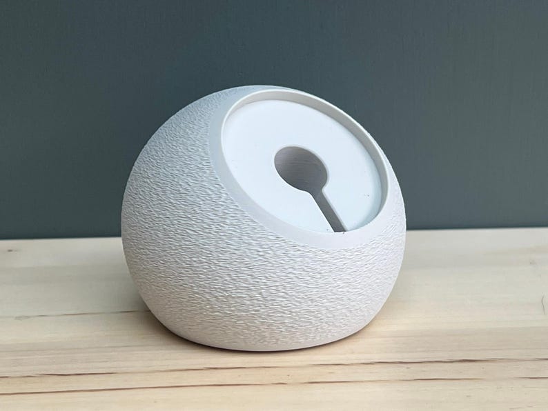 May include: A spherical white charging dock with a textured, matte exterior surface. The top features a smooth, recessed circular area with a central hole and a vertical slot extending downward, designed to hold a charging cable securely in place.