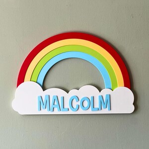 May include: A colorful rainbow-shaped wall decoration with the name "MALCOLM" in blue letters. The rainbow features red, yellow, green, and blue arches above white cloud cutouts. This decorative piece is designed for a child's room.