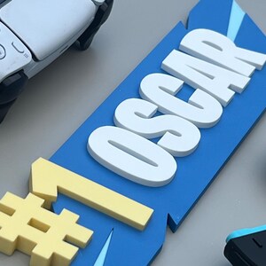 May include: A blue sign with the name "OSCAR" in white, 3D letters, and a yellow number one symbol. A white and black game controller is in the background, along with other game controllers.