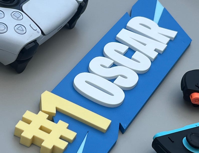May include: A blue sign with the name "OSCAR" in white, 3D letters, and a yellow number one symbol. A white and black game controller is in the background, along with other game controllers.