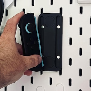 May include: A black and light blue video game controller being placed into a black wall mount. The wall mount is attached to a white pegboard. The controller has black buttons and a light blue accent.