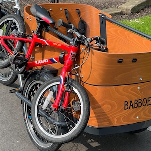May include: A red Wiggins bicycle is attached to a wooden cargo bike with the word BABBOE printed on the side. The cargo bike has a black seat and a black storage bag. The bicycle has a blue and white stripe design.