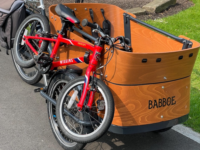 May include: A red Wiggins bicycle is attached to a wooden cargo bike with the word BABBOE printed on the side. The cargo bike has a black seat and a black storage bag. The bicycle has a blue and white stripe design.