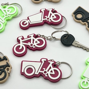 May include: A collection of colorful keychains featuring a bicycle with a cargo box. The keychains are made of rubber and have a white outline. Some of the keychains are green, red, and black.