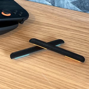 May include: Two black gaming controller accessories with illuminated ends, one blue and one orange, are crossed on a wooden surface. A portion of a black gaming controller with orange accents is visible in the background.
