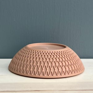 May include: A dusty rose-coloured bowl with a rounded shape and a textured exterior featuring a repeating diamond lattice pattern. The bowl has a smooth, flat circular base at the top and a wide, circular rim at the bottom, presenting a clean, uniform finish.