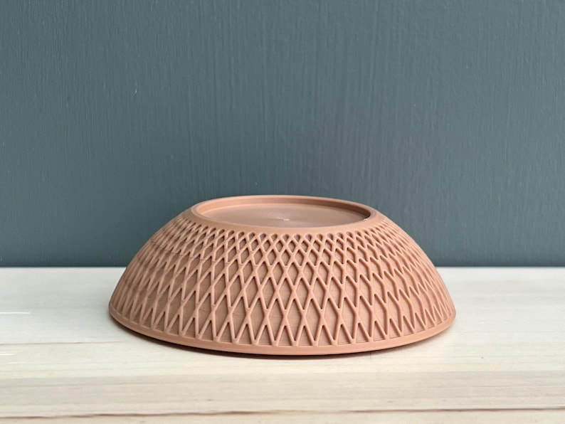 May include: A dusty rose-coloured bowl with a rounded shape and a textured exterior featuring a repeating diamond lattice pattern. The bowl has a smooth, flat circular base at the top and a wide, circular rim at the bottom, presenting a clean, uniform finish.