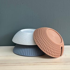 May include: Three stacked bowls in different colours and textures. The top bowl is white with a wavy surface, the middle bowl is dark grey with vertical ridges, and the front bowl is terracotta with a geometric diamond-shaped lattice pattern.