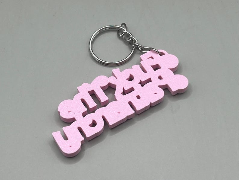 May include: A pink keychain with the word "sorry" in a stylized font. The keychain is attached to a silver metal ring. The background is a solid gray color.