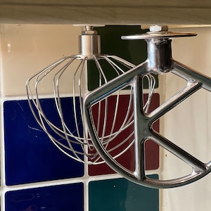 May include: Two silver kitchen mixer attachments hanging from a shelf. The attachment on the left is a wire whisk. The attachment on the right is a flat paddle.