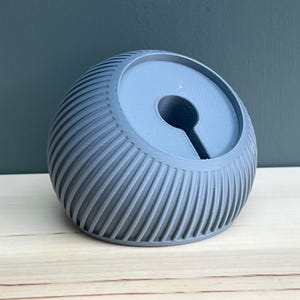 May include: A matte grey, spherical, 3D-printed stand featuring a ribbed, textured exterior. The top surface is flat and circular with a central, keyhole-shaped cutout designed to hold a device or cable, showcasing a layered construction.