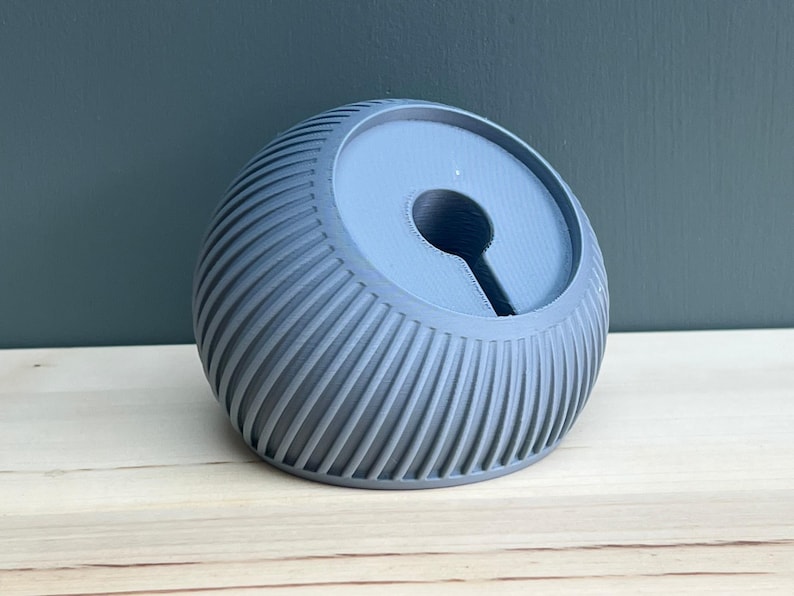 May include: A matte grey, spherical, 3D-printed stand featuring a ribbed, textured exterior. The top surface is flat and circular with a central, keyhole-shaped cutout designed to hold a device or cable, showcasing a layered construction.