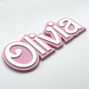 Puede incluir: Un letrero de nombre 3D rosa y blanco que dice "Olivia". Las letras son en relieve y tienen un contorno blanco.
