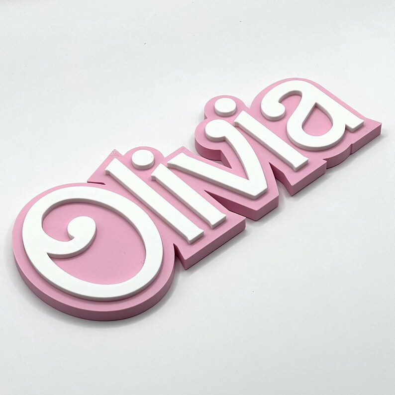 Puede incluir: Un letrero de nombre 3D rosa y blanco que dice "Olivia". Las letras son en relieve y tienen un contorno blanco.