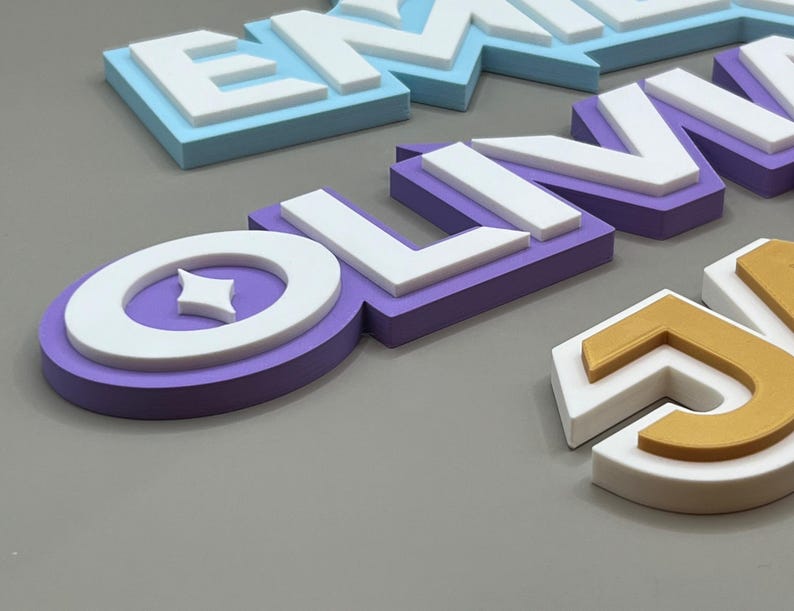 May include: Close-up of colorful, layered wooden letters spelling out names. The letters are in various colors, including blue, purple, and gold, with white accents. The letters are arranged on a gray surface.