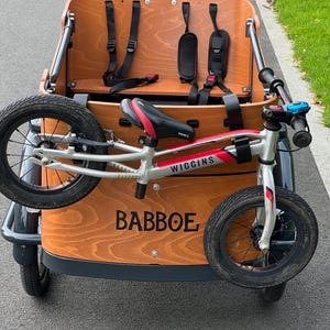 May include: A wooden cargo bike with a silver and red Wiggins balance bike attached. The cargo bike has a brown wooden box with black straps and a black seat. The balance bike has black tires and a red and black seat.