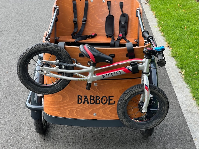 May include: A wooden cargo bike with a silver and red Wiggins balance bike attached. The cargo bike has a brown wooden box with black straps and a black seat. The balance bike has black tires and a red and black seat.