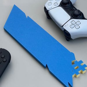 May include: A blue foam sword prop with a hashtag-shaped hilt, alongside a white and black game controller and a black and orange game controller. The sword has jagged edges and is made of solid blue foam.