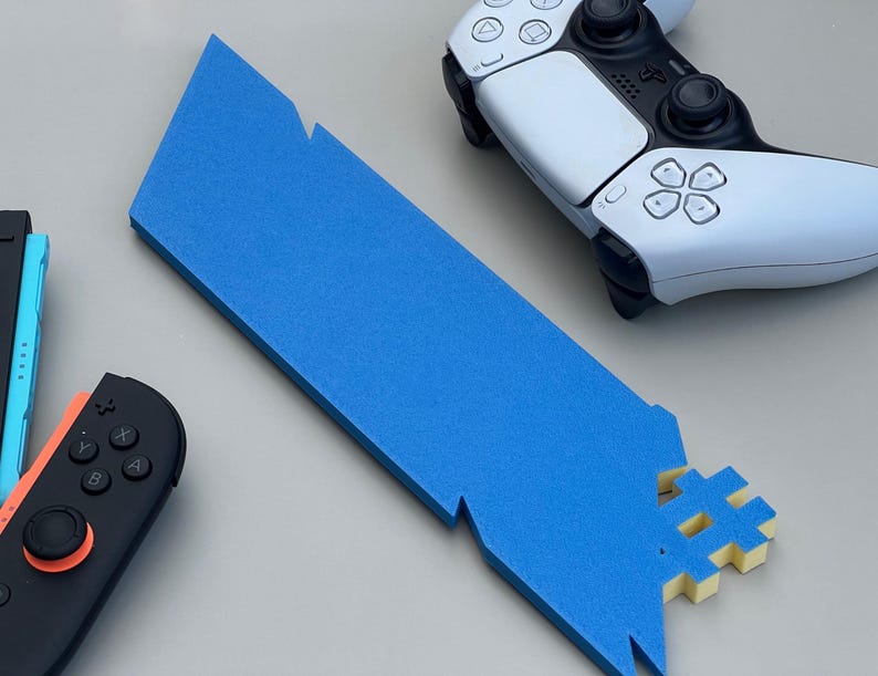 May include: A blue foam sword prop with a hashtag-shaped hilt, alongside a white and black game controller and a black and orange game controller. The sword has jagged edges and is made of solid blue foam.