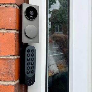 May include: A gray doorbell with a camera and a keypad. The doorbell is mounted on a brick wall. The keypad has numbers 1 through 9, 0, and a checkmark.
