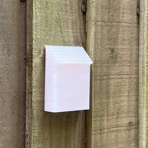 May include: White plastic birdhouse with a small roof, mounted on a wooden fence.