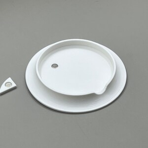 May include: A white, circular lid with a small, embedded magnet, resting on a larger, matching base. A separate, white, triangular piece with a magnet is positioned to the left. The items are on a gray surface.