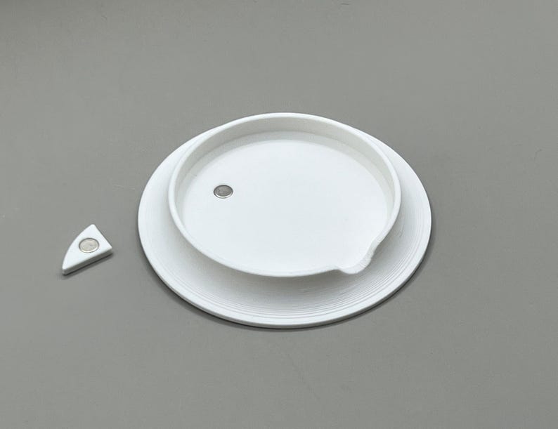 May include: A white, circular lid with a small, embedded magnet, resting on a larger, matching base. A separate, white, triangular piece with a magnet is positioned to the left. The items are on a gray surface.