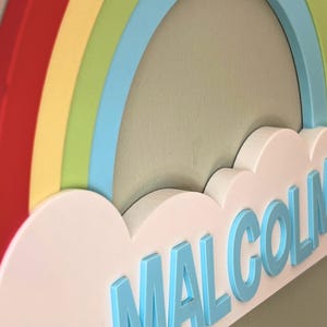 May include: A colorful rainbow wall decor with the name "Malcolm" in light blue letters. The rainbow features red, yellow, green, and blue arches. The name is placed on a white cloud-shaped base.