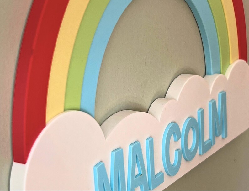 May include: A colorful rainbow wall decor with the name "Malcolm" in light blue letters. The rainbow features red, yellow, green, and blue arches. The name is placed on a white cloud-shaped base.