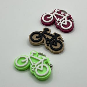 May include: Three bike-shaped keychains in different colors: maroon, beige, and lime green. Each keychain features a white bike silhouette.