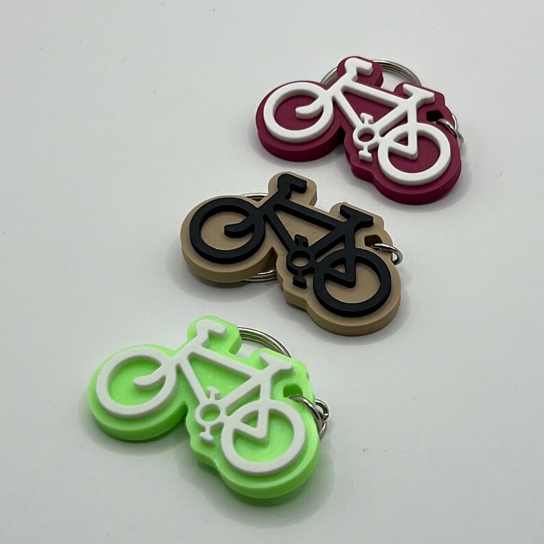 May include: Three bike-shaped keychains in different colors: maroon, beige, and lime green. Each keychain features a white bike silhouette.