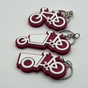 May include: Three pink and white bicycle keychains with silver key rings. The keychains are shaped like bicycles with a cargo box on the back.