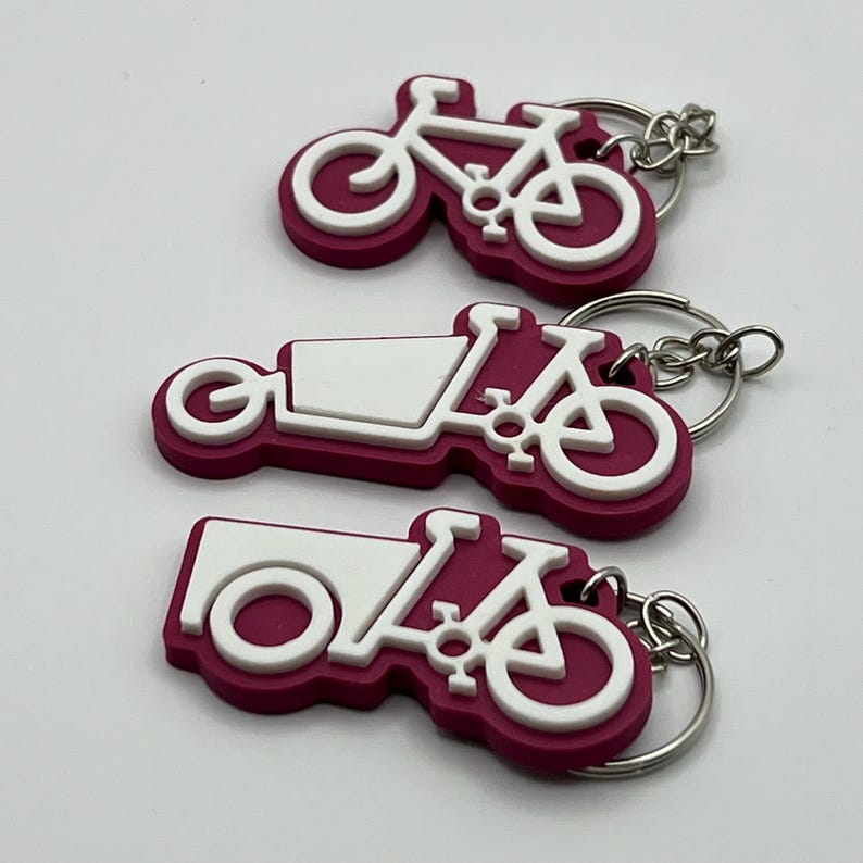 May include: Three pink and white bicycle keychains with silver key rings. The keychains are shaped like bicycles with a cargo box on the back.
