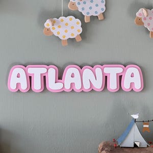 May include: Pink wooden name sign with the word "ATLANTA" in white raised letters. The sign is hanging on a wall with a green background.