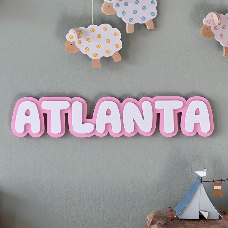 May include: Pink wooden name sign with the word "ATLANTA" in white raised letters. The sign is hanging on a wall with a green background.