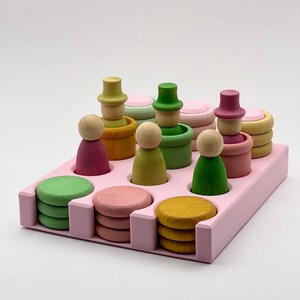 May include: A pink wooden tray with 12 compartments holds a set of colorful wooden stacking toys. The set includes green, yellow, pink, and brown wooden figures with hats, and a set of colorful wooden circles.