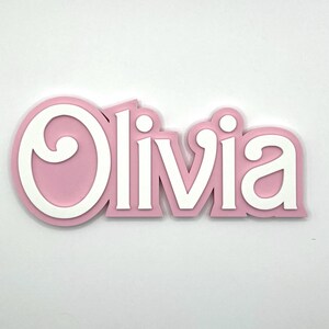 Puede incluir: Un letrero de nombre de madera rosa y blanco con el nombre "Olivia" en una fuente decorativa.