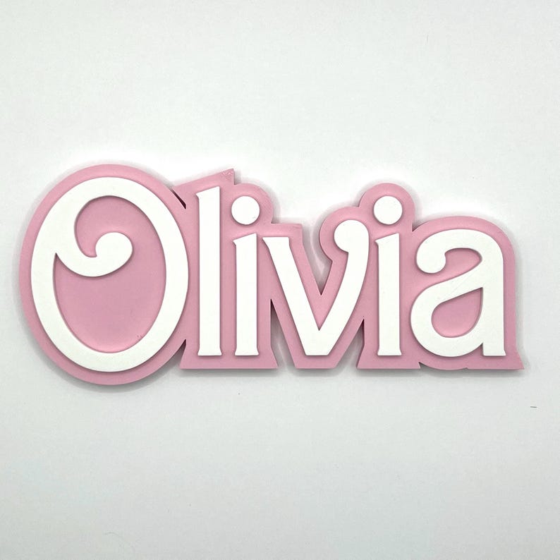 Puede incluir: Un letrero de nombre de madera rosa y blanco con el nombre "Olivia" en una fuente decorativa.