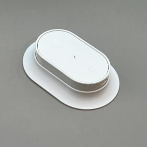 Pu&ograve; includere: Un dispositivo bianco di forma ovale con un piccolo punto nero sulla parte superiore, appoggiato su una base ovale bianca abbinata. Il dispositivo ha una fascia argentata attorno al centro. Il design &egrave; minimalista e moderno.