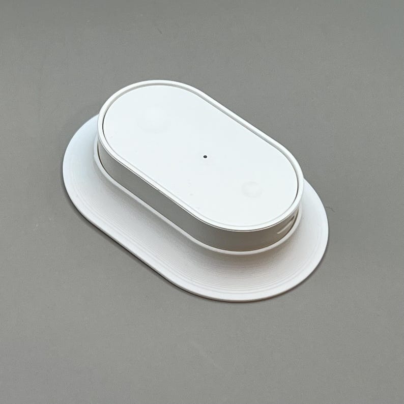 Pu&ograve; includere: Un dispositivo bianco di forma ovale con un piccolo punto nero sulla parte superiore, appoggiato su una base ovale bianca abbinata. Il dispositivo ha una fascia argentata attorno al centro. Il design &egrave; minimalista e moderno.