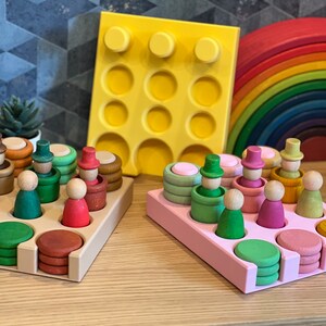 May include: Two wooden trays with different colored bases, one beige and one pink, hold various wooden toys. The beige tray has a set of small wooden people with hats, and the pink tray has a set of stacking rings and small wooden people.