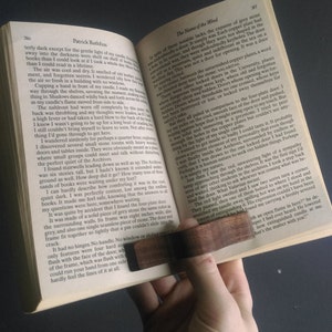 May include: A close-up of an open book with a wooden bookmark. The book is titled "The Name of the Wind" by Patrick Rothfuss. The text on the page describes a large, grey stone door with no hinges or handle.